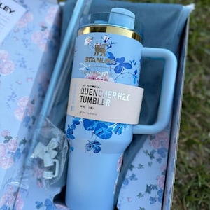 May include: A light blue Stanley LoveShackFancy Quencher H2.0 tumbler with a floral pattern, a gold-tone rim, and a handle. The tumbler features a paper band labeled The Flowstate Quencher H2.0 Tumbler 40 oz, accompanied by a straw and accessories.