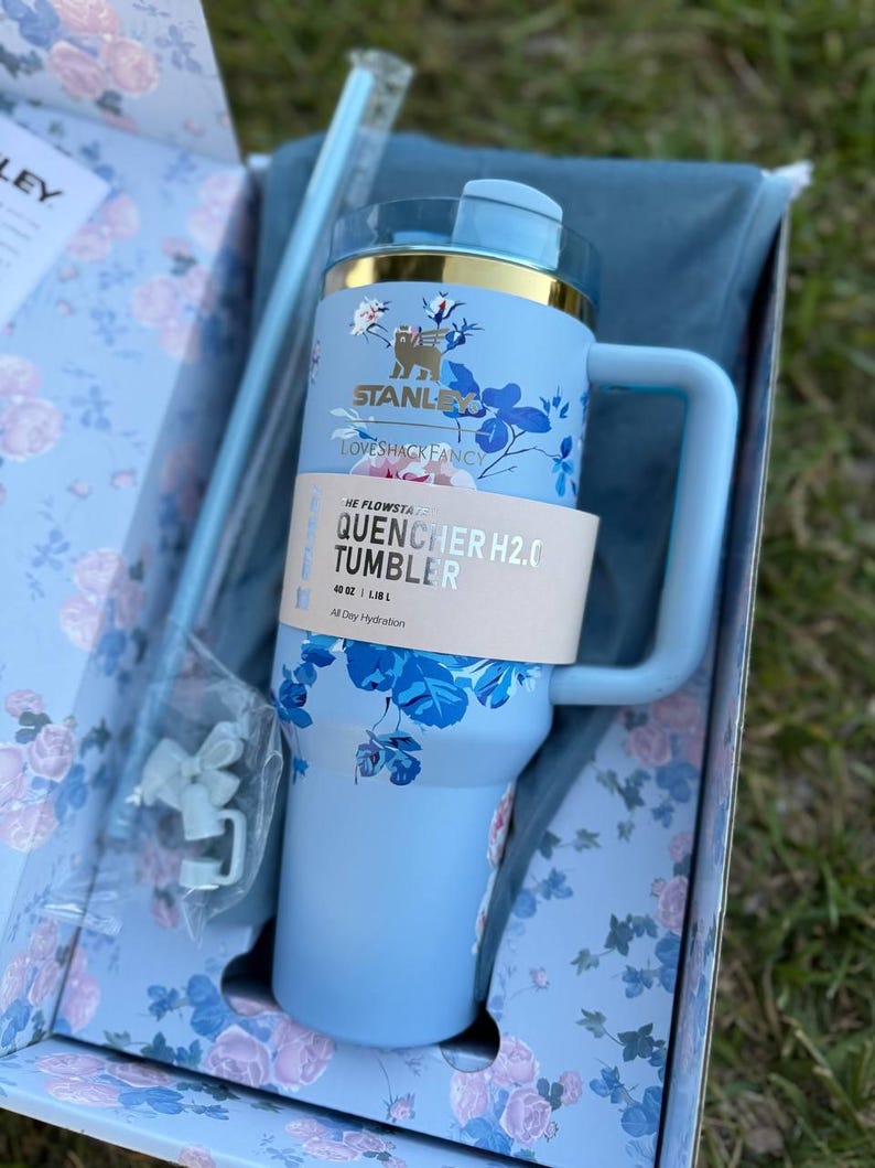 May include: A light blue Stanley LoveShackFancy Quencher H2.0 tumbler with a floral pattern, a gold-tone rim, and a handle. The tumbler features a paper band labeled The Flowstate Quencher H2.0 Tumbler 40 oz, accompanied by a straw and accessories.