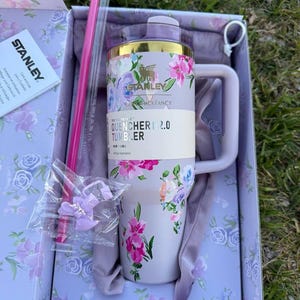 May include: A light purple Stanley Quencher H2.0 tumbler with a floral pattern, gold rim, and handle. It includes a pink straw, a small bow accessory in a plastic bag, and a care instruction booklet, all packaged in a matching floral box.