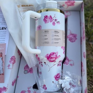 Op de afbeelding: Een witte Stanley-beker met een roze rozenpatroon, een goudkleurige rand en een wit handvat. De beker zit in een bijpassende bloemendoos met een doorzichtig deksel, een wit rietje en een klein wit accessoire in de verpakking.