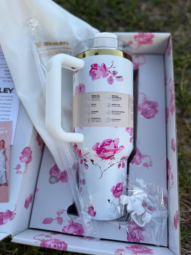 Op de afbeelding: Een witte Stanley-beker met een roze rozenpatroon, een goudkleurige rand en een wit handvat. De beker zit in een bijpassende bloemendoos met een doorzichtig deksel, een wit rietje en een klein wit accessoire in de verpakking.