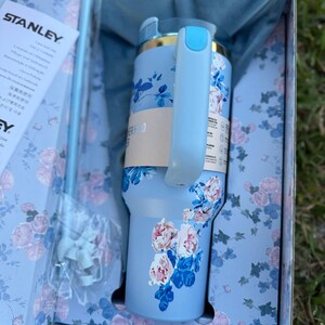 May include: A light blue Stanley tumbler with a floral pattern of pink and white roses and dark blue leaves. The tumbler features a light blue handle, a matching straw, and a blue fabric pouch, all presented in a matching floral gift box.