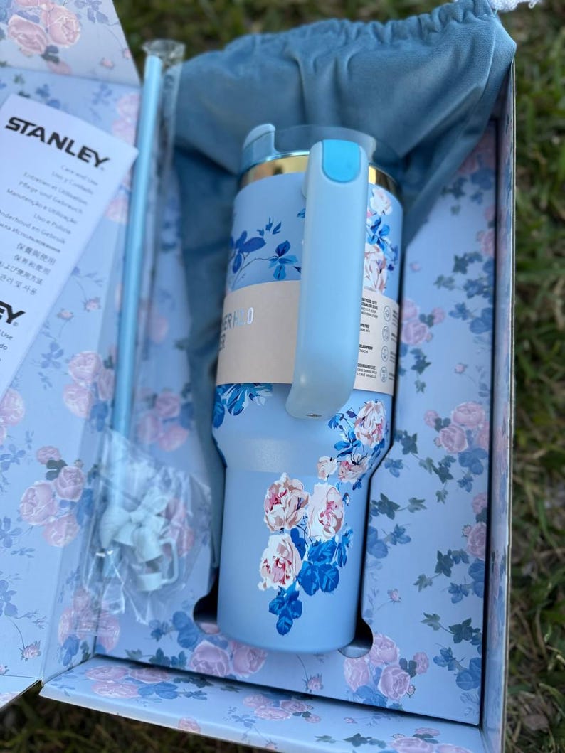 May include: A light blue Stanley tumbler with a floral pattern of pink and white roses and dark blue leaves. The tumbler features a light blue handle, a matching straw, and a blue fabric pouch, all presented in a matching floral gift box.