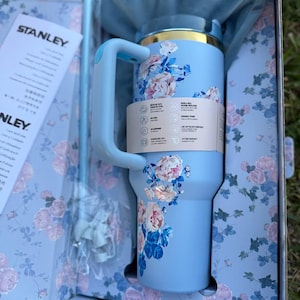 May include: A light blue Stanley tumbler with a floral pattern featuring pink roses and green leaves. The tumbler has a handle, a gold-tone rim, and a matching straw. It is presented in a matching floral box with a blue fabric pouch and instruction manual.