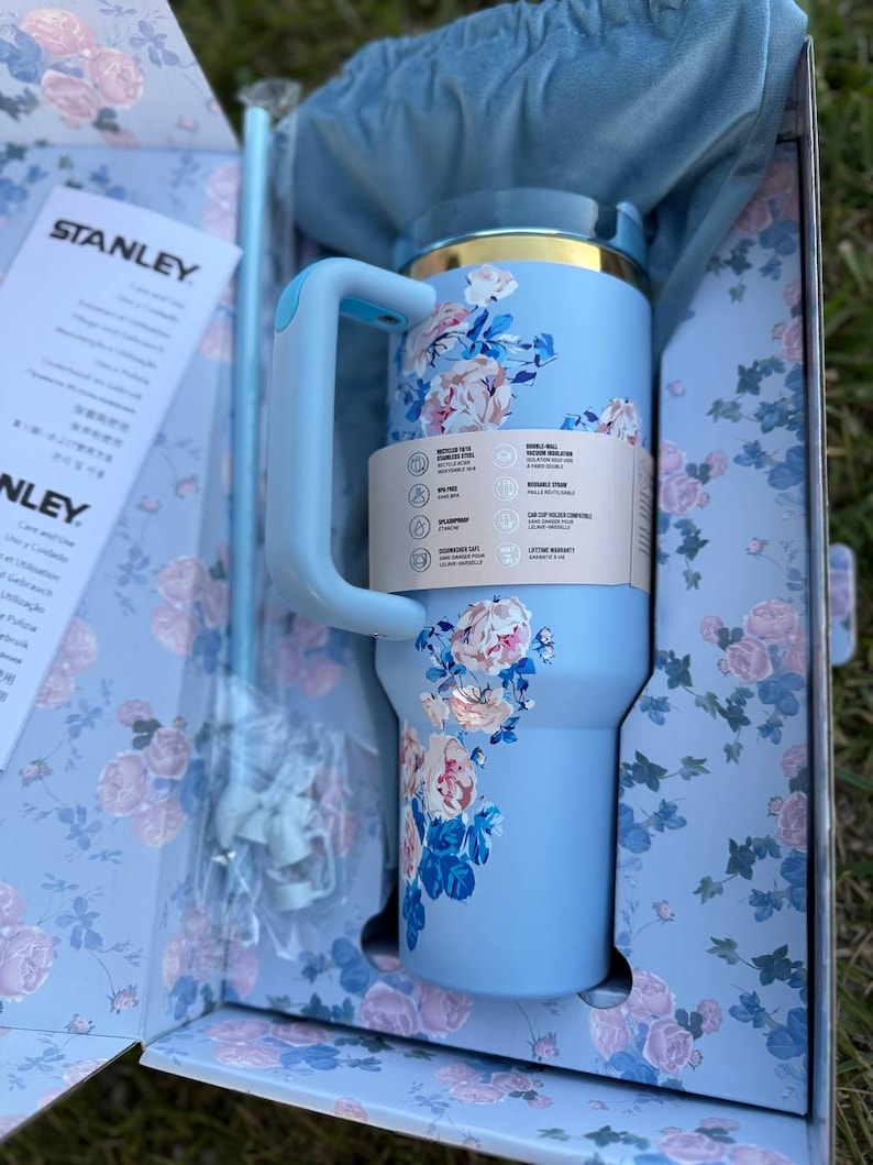 May include: A light blue Stanley tumbler with a floral pattern featuring pink roses and green leaves. The tumbler has a handle, a gold-tone rim, and a matching straw. It is presented in a matching floral box with a blue fabric pouch and instruction manual.