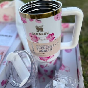 Op de afbeelding: Een witte Stanley Quencher H2.0 FlowState beker met een roze bloemenpatroon, een goudkleurige rand en een witte handgreep. De beker heeft een papieren band met de tekst Stanley Quencher H2.0 Tumbler 1,18 L, een doorzichtig deksel en witte accessoires.