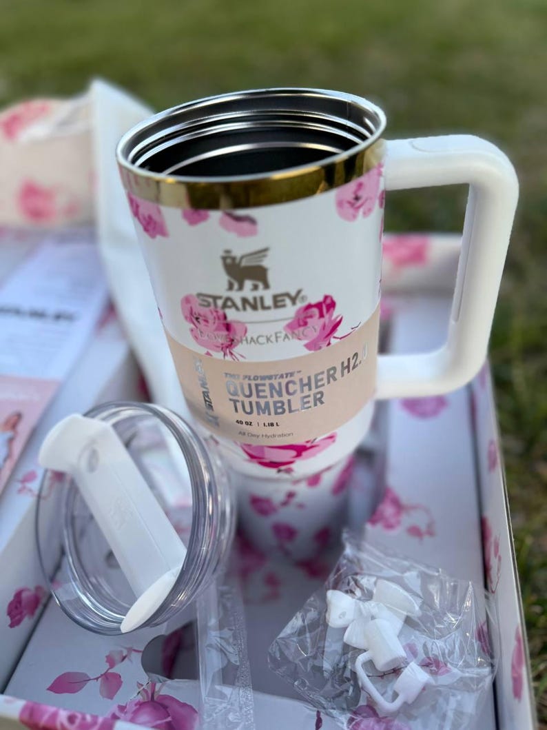 Op de afbeelding: Een witte Stanley Quencher H2.0 FlowState beker met een roze bloemenpatroon, een goudkleurige rand en een witte handgreep. De beker heeft een papieren band met de tekst Stanley Quencher H2.0 Tumbler 1,18 L, een doorzichtig deksel en witte accessoires.