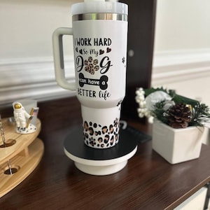May include: A white stainless steel tumbler with a handle and a lid, featuring black text that reads I WORK HARD So My DOG can have a BETTER life, with a leopard print paw print and a leopard print pattern around the base of the tumbler.