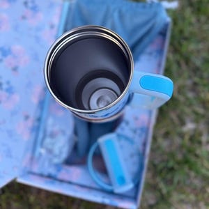 May include: A stainless steel tumbler with a light blue handle and a matching lid. The tumbler features a cylindrical interior and is positioned within a floral patterned box. The handle and lid include bright blue accents on the exterior.