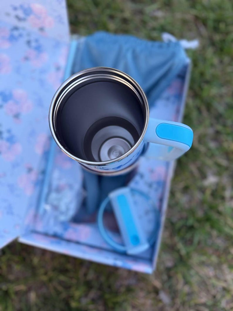 May include: A stainless steel tumbler with a light blue handle and a matching lid. The tumbler features a cylindrical interior and is positioned within a floral patterned box. The handle and lid include bright blue accents on the exterior.