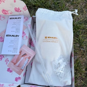 Op de afbeelding: Een witte stoffen tas met trekkoord en de gouden Stanley en LoveShackFancy logo's, geplaatst in een doos met bloemenpatroon. Inbegrepen zijn een doorzichtig plastic rietje, een zakje met witte onderdelen en twee informatiebladen.