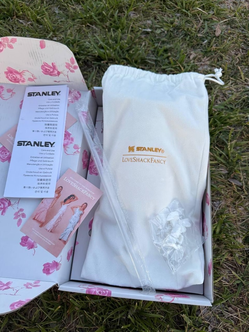 Op de afbeelding: Een witte stoffen tas met trekkoord en de gouden Stanley en LoveShackFancy logo's, geplaatst in een doos met bloemenpatroon. Inbegrepen zijn een doorzichtig plastic rietje, een zakje met witte onderdelen en twee informatiebladen.