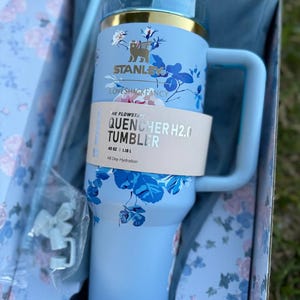 May include: A light blue Stanley Quencher H2.0 Flowstate tumbler with a 40 oz capacity, featuring a floral pattern, a gold-tone rim, and a side handle. A paper band displays the text The Flowstate Quencher H2.0 Tumbler 40 oz | 1.18 L All Day Hydration.