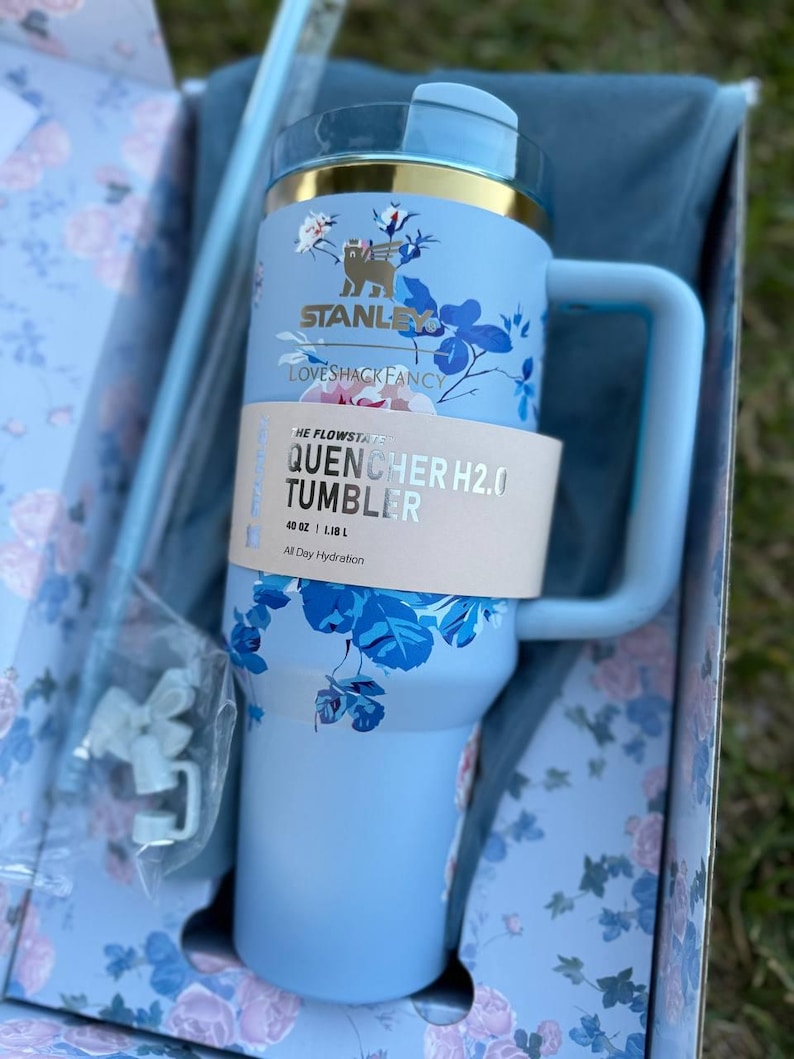 May include: A light blue Stanley Quencher H2.0 Flowstate tumbler with a 40 oz capacity, featuring a floral pattern, a gold-tone rim, and a side handle. A paper band displays the text The Flowstate Quencher H2.0 Tumbler 40 oz | 1.18 L All Day Hydration.