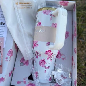 Op de afbeelding: Een witte Stanley Quencher H2.0 beker met een roze rozenpatroon, een goudkleurige rand en een witte handgreep. De beker zit in een bijpassende bloemendoos met een witte stofzak, een doorzichtig rietje en een klein wit strikje als accessoire.