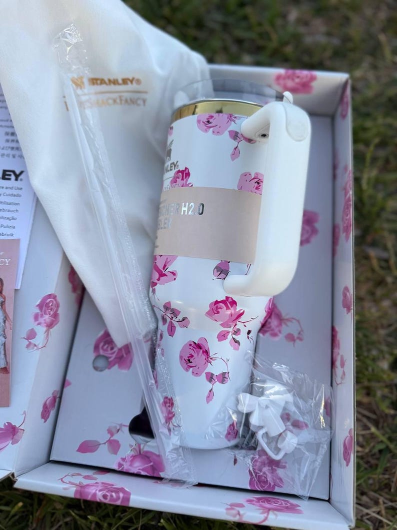 Op de afbeelding: Een witte Stanley Quencher H2.0 beker met een roze rozenpatroon, een goudkleurige rand en een witte handgreep. De beker zit in een bijpassende bloemendoos met een witte stofzak, een doorzichtig rietje en een klein wit strikje als accessoire.
