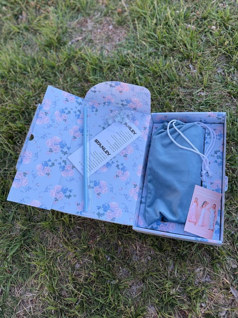 May include: A rectangular gift box with a light blue floral pattern containing a light blue straw, a white Stanley brand information card, a blue drawstring pouch, and a small rectangular card featuring images of a person in a long dress.