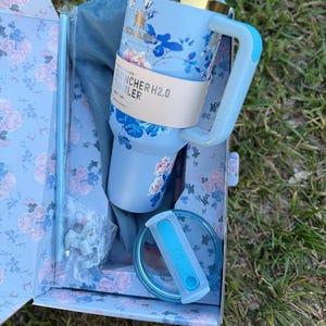 May include: A light blue Stanley Quencher H2.0 tumbler with a floral pattern, a gold-tone rim, and a matching handle. The tumbler includes a clear lid with a blue insert, a straw, and a blue drawstring bag, all packaged in a floral box.