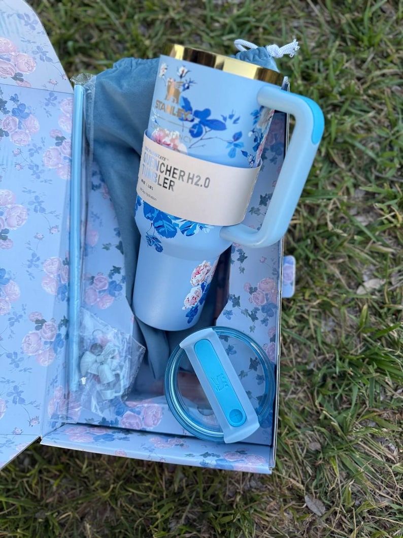 May include: A light blue Stanley Quencher H2.0 tumbler with a floral pattern, a gold-tone rim, and a matching handle. The tumbler includes a clear lid with a blue insert, a straw, and a blue drawstring bag, all packaged in a floral box.