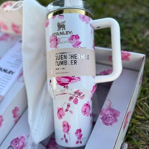 Op de afbeelding: Een witte Stanley Quencher H2.0 beker met een roze rozenpatroon, een goudkleurige rand en een wit handvat. De beker heeft een beige papieren band met de tekst Stanley, LoveShackFancy, The FlowState, Quencher H2.0 Tumbler, 1,18 L.