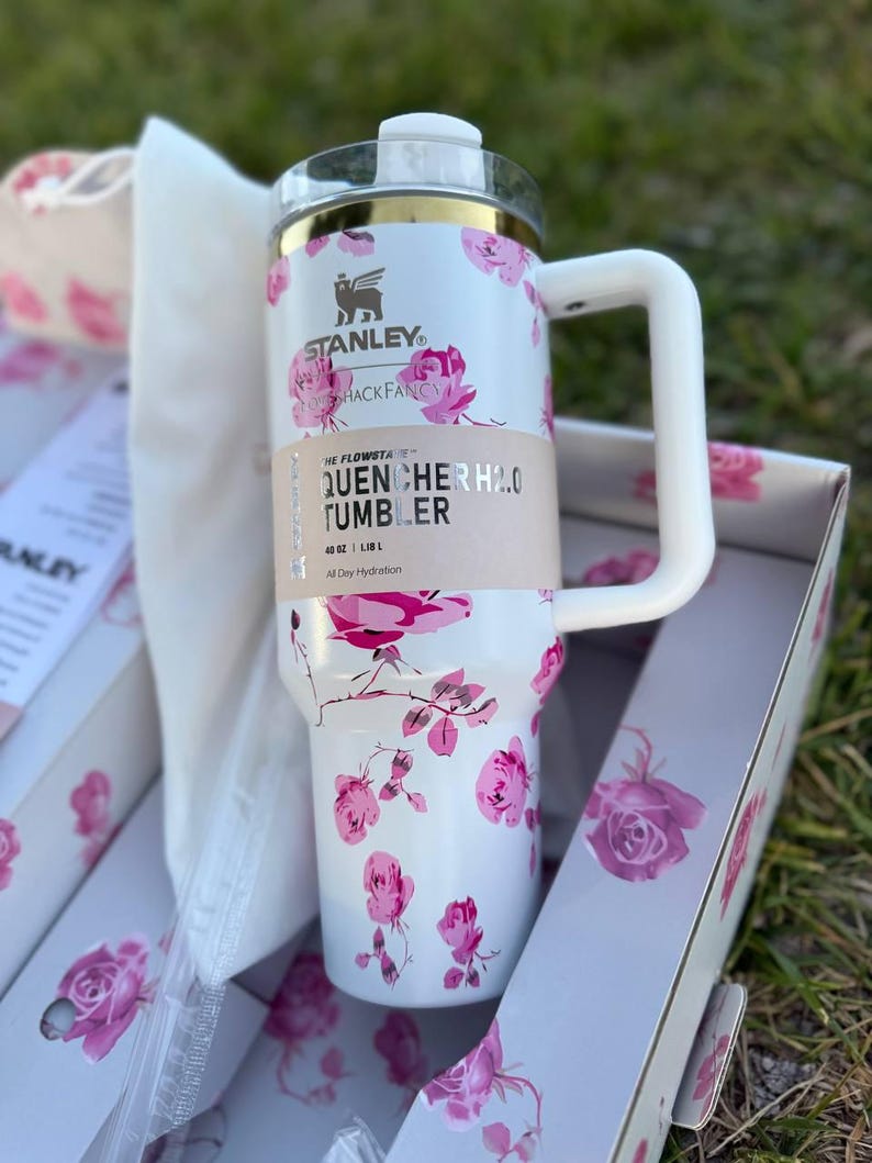 Op de afbeelding: Een witte Stanley Quencher H2.0 beker met een roze rozenpatroon, een goudkleurige rand en een wit handvat. De beker heeft een beige papieren band met de tekst Stanley, LoveShackFancy, The FlowState, Quencher H2.0 Tumbler, 1,18 L.