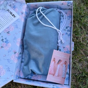 May include: A light blue drawstring pouch rests inside a rectangular box featuring a floral pattern of pink roses and blue leaves. The box contains a white Stanley care instruction card and a rectangular LoveShackFancy brand card.