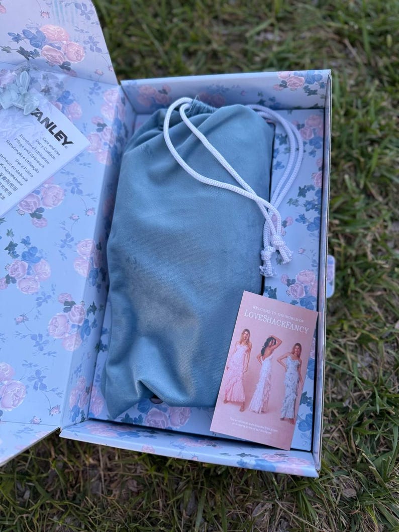 May include: A light blue drawstring pouch rests inside a rectangular box featuring a floral pattern of pink roses and blue leaves. The box contains a white Stanley care instruction card and a rectangular LoveShackFancy brand card.