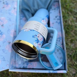 May include: A light blue insulated tumbler with a floral pattern, a gold-tone rim, and a matching handle. The tumbler includes a clear lid with a blue sliding closure and is presented in a matching floral-patterned box with a blue fabric pouch.