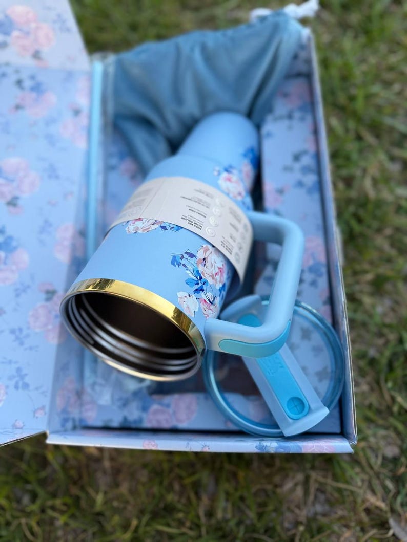 May include: A light blue insulated tumbler with a floral pattern, a gold-tone rim, and a matching handle. The tumbler includes a clear lid with a blue sliding closure and is presented in a matching floral-patterned box with a blue fabric pouch.