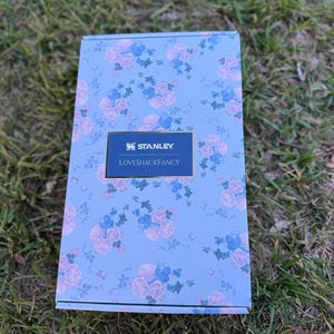 May include: A rectangular cardboard box with a light blue background featuring a repeating pattern of pink roses and blue flowers. A centered dark blue rectangular label displays the white text Stanley and LoveShackFancy in a serif font.