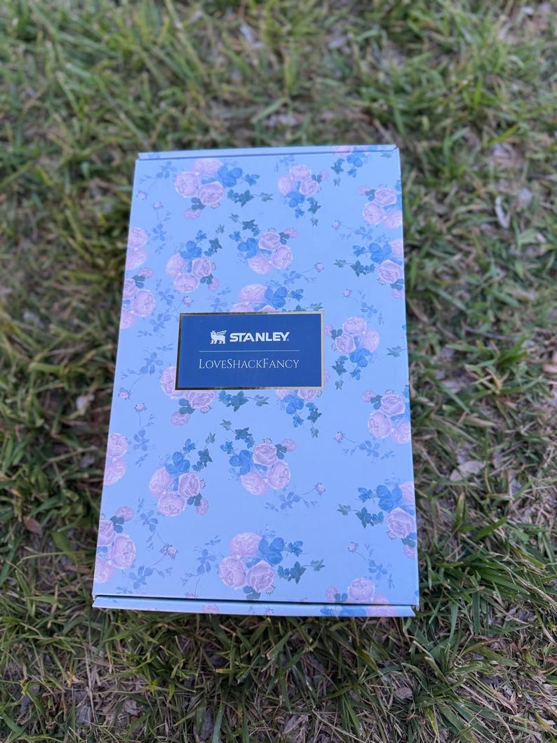 May include: A rectangular cardboard box with a light blue background featuring a repeating pattern of pink roses and blue flowers. A centered dark blue rectangular label displays the white text Stanley and LoveShackFancy in a serif font.