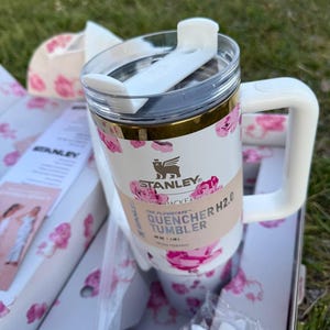 Op de afbeelding: Een witte Stanley Quencher H2.0 beker met een roze bloemenpatroon, een doorzichtig deksel met een witte draaibare sluiting en een wit handvat. De beker is voorzien van een papieren wikkel met de tekst Stanley Quencher H2.0 Tumbler.