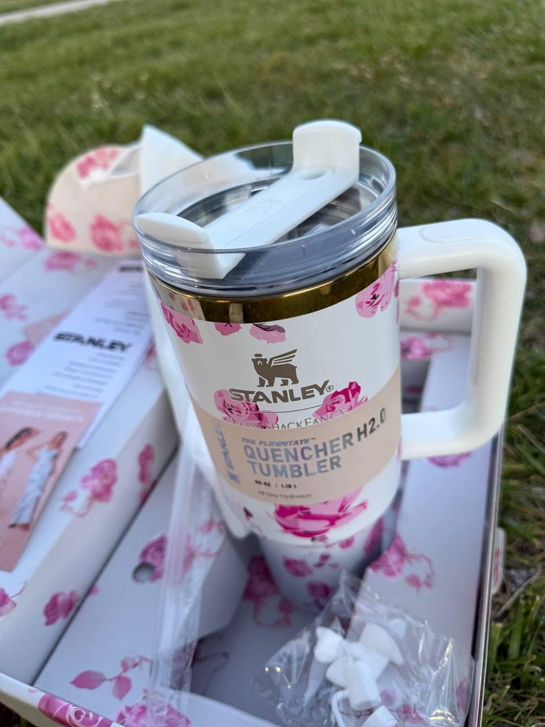 Op de afbeelding: Een witte Stanley Quencher H2.0 beker met een roze bloemenpatroon, een doorzichtig deksel met een witte draaibare sluiting en een wit handvat. De beker is voorzien van een papieren wikkel met de tekst Stanley Quencher H2.0 Tumbler.