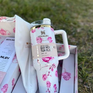 Op de afbeelding: Een witte Stanley Quencher H2.0 beker met een roze rozenpatroon, een goudkleurige rand en een wit handvat. De beker heeft een label met de tekst The Flowstate Quencher H2.0 Tumbler 40 oz / 1,18 L, vergezeld van een bijpassende doos en tas.