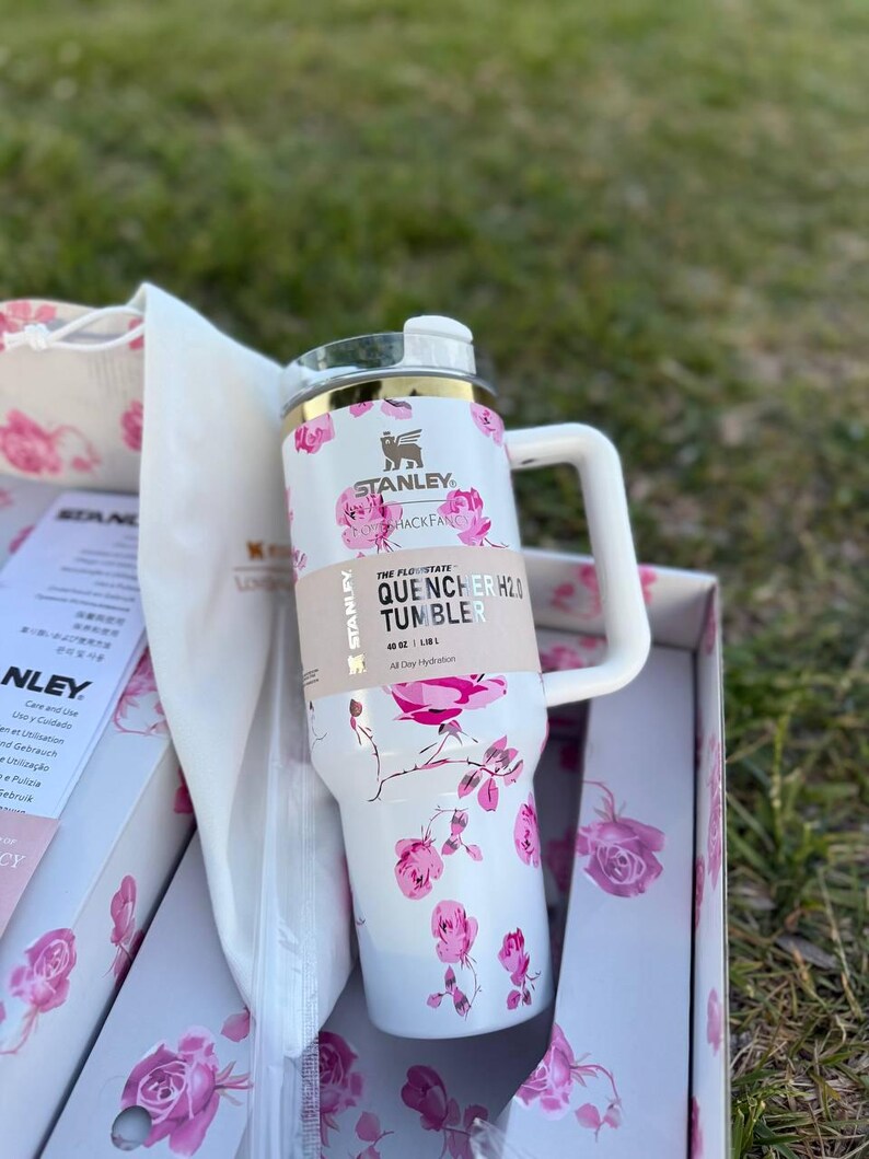 Op de afbeelding: Een witte Stanley Quencher H2.0 beker met een roze rozenpatroon, een goudkleurige rand en een wit handvat. De beker heeft een label met de tekst The Flowstate Quencher H2.0 Tumbler 40 oz / 1,18 L, vergezeld van een bijpassende doos en tas.