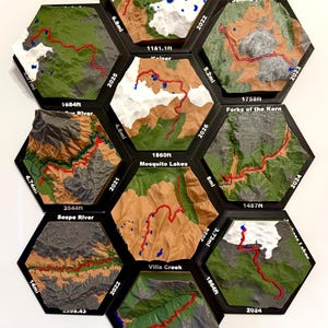 May include: A collection of nine hexagonal 3D topographic map tiles arranged in a cluster. Each tile features a raised terrain model with a red line indicating a hiking trail, blue spots for water, and printed text showing location names, years, and distances in miles and feet.
