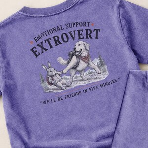 May include: A purple t-shirt featuring a graphic print of a dog and a rabbit holding hands. The text reads EMOTIONAL SUPPORT EXTROVERT and WE'LL BE FRIENDS IN FIVE MINUTES. The illustration includes a dog with a backpack and a rabbit in a hoodie.