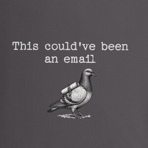 May include: A dark gray fabric surface featuring a white graphic illustration of a pigeon carrying a rolled scroll on its back. Above the bird, the text reads, "This could've been an email" in a white, typewriter-style font. The design is centered.