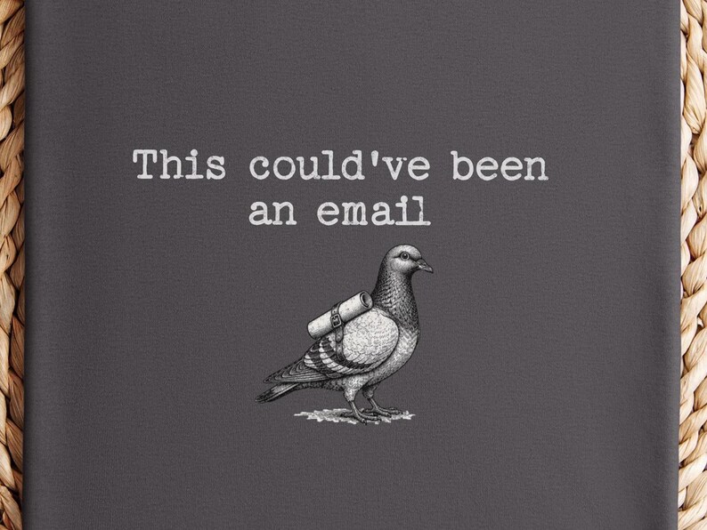 May include: A dark gray fabric surface featuring a white graphic illustration of a pigeon carrying a rolled scroll on its back. Above the bird, the text reads, "This could've been an email" in a white, typewriter-style font. The design is centered.