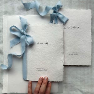 May include: Two rectangular off-white handmade paper cards with deckled edges, each tied with a light blue frayed silk ribbon. One card reads to my wife.. on our wedding day 23rd May 2026, and the other reads to my husband.. on our wedding day 23rd May 2026.