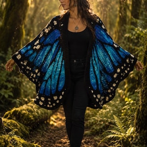 May include: A crochet cardigan featuring a vibrant blue and black butterfly wing pattern with white accents along the edges. The garment is worn over a black tank top and black pants, with a pendant necklace and black ankle boots completing the look.
