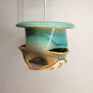 May include: A hanging ceramic planter with a glossy turquoise and teal glaze on the upper section and a natural tan base. The vessel features multiple side openings for planting and a single white rope cord extending from the top centre.