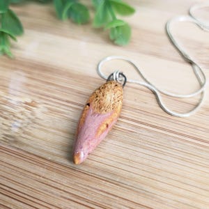May include: A pendant necklace with a distinctive, elongated pendant. The pendant combines natural wood tones with a soft pink colour, suspended from a delicate silver chain. The necklace is set against a wooden surface.