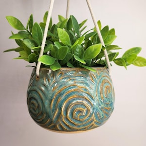 May include: A round ceramic hanging planter with a teal and brown swirled glaze pattern. The pot features a textured surface with carved spiral designs and is suspended by three thick white cotton cords attached to the rim of the vessel.