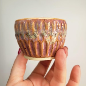 May include: A small, handmade ceramic bowl with a textured, ridged exterior. The bowl features a blend of purple, gold, and green hues. The rim is a warm gold colour, and the base is a light beige. The bowl is held in a hand.
