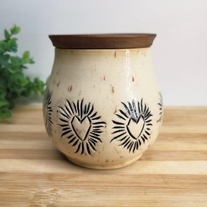 May include: A beige ceramic jar with a brown lid. The jar features a repeating pattern of black heart designs with radiating lines. The jar is sitting on a wooden surface.