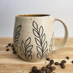 May include: A cream-coloured ceramic mug with a speckled finish and a black hand-drawn leaf pattern. The mug features a rounded body, a curved handle, and a smooth glazed surface. The design consists of vertical stems with pointed leaves.
