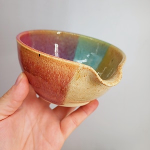 May include: A handmade ceramic bowl with a colourful, striped design. The bowl features a unique spout and is held in a hand. The colours include red, purple, blue, and yellow.