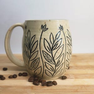 May include: A cream-coloured ceramic mug with a speckled finish and a dark, hand-carved botanical pattern featuring vertical stems and leaves. The mug has a rounded body and a curved handle, with several roasted coffee beans scattered nearby.