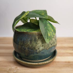 Peut inclure: Un pot de fleurs rond en céramique avec un émail réactif présentant des nuances mélangées de bleu profond, de vert forêt et de brun terreux. Le pot repose sur une soucoupe circulaire assortie et contient une petite plante aux feuilles panachées.